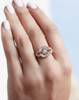 A 14k rose gold ring with a geometric, art deco style diamond-like pattern, set with one center brilliant cut white diamond and four smaller ones, worn on a hand with pale pink manicured nails, set against a soft, out-of-focus light blue and white background.