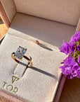A gold ring with a radiant cut white diamond displayed on a beige velvet ring box, set on a wooden surface with purple flowers nearby.