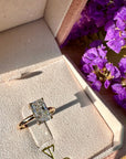 A gold ring with a radiant cut lab grown diamond displayed inside a pink velvet jewelry box with the brand name TOR printed on the inside lid. Purple flowers are visible on the right side.