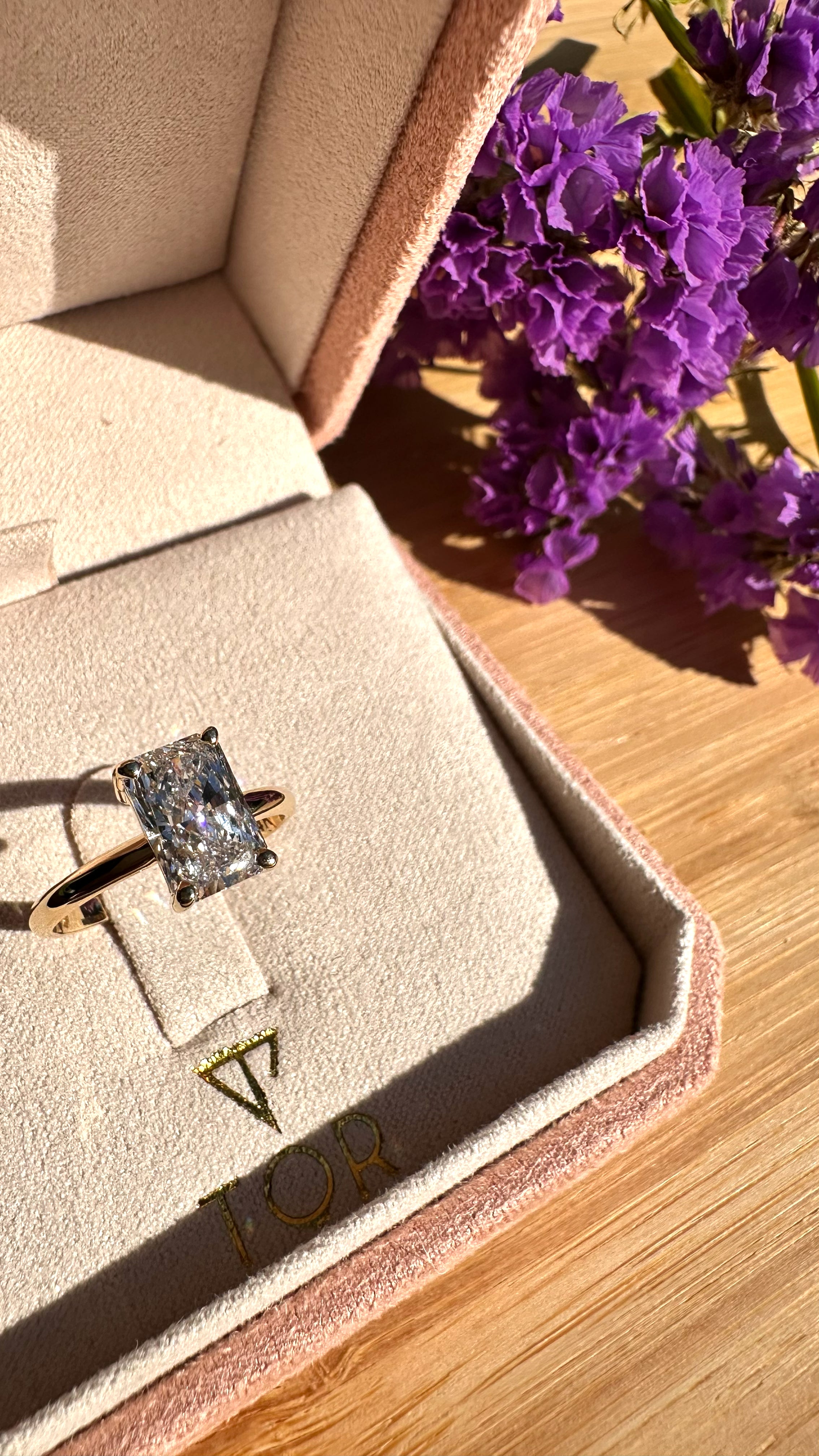 A gold ring with a radiant cut white diamond displayed inside an open beige jewelry box on a wooden surface, with purple flowers in the background.