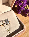 A gold ring with a radiant cut white diamond displayed inside an open beige jewelry box on a wooden surface, with purple flowers in the background.