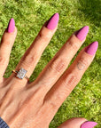A close-up of a hand with long pink manicured nails wearing a radiant cut diamond engagement ring on a grassy lawn.
