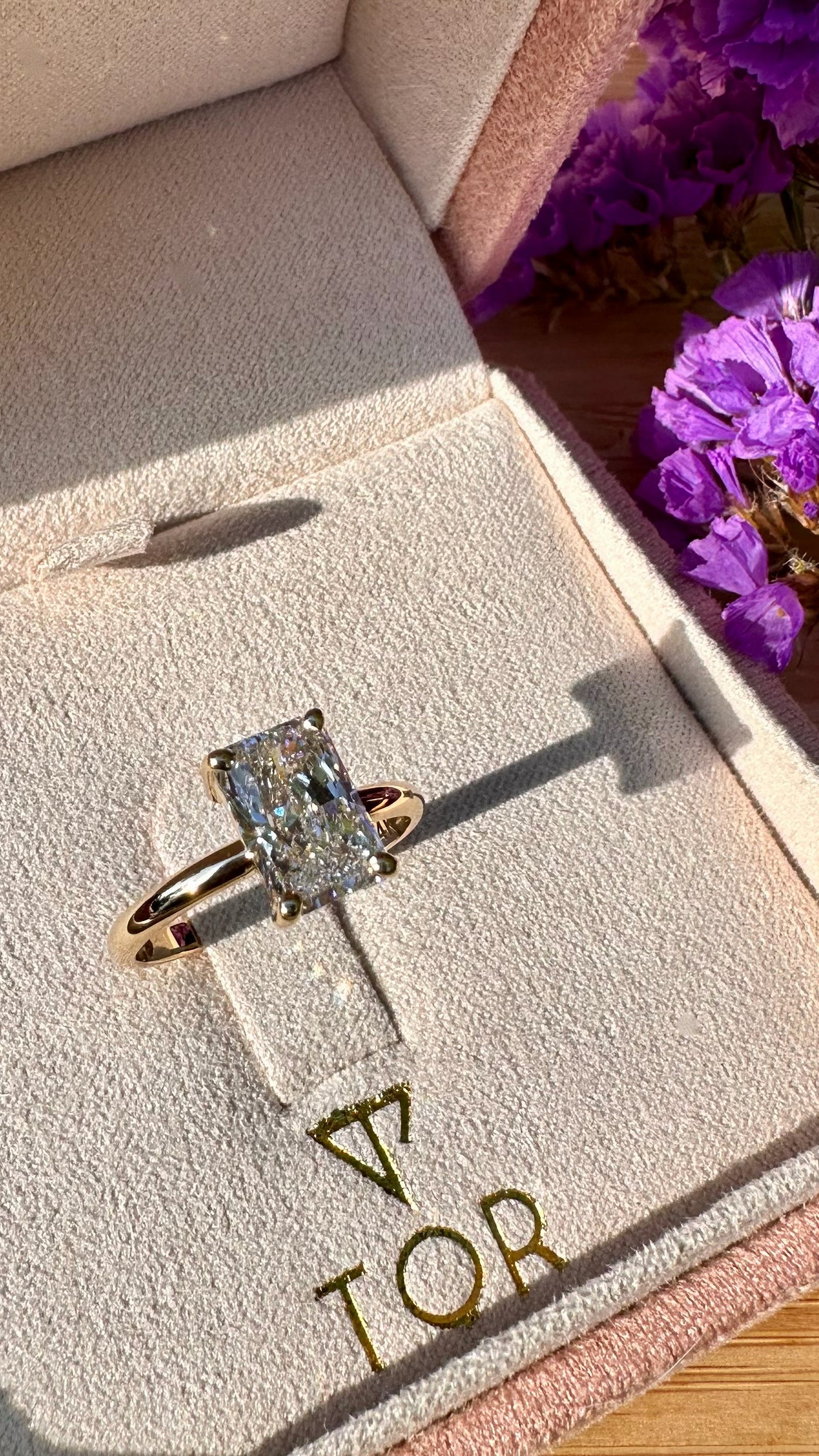 A radiant cut white diamond ring with a gold band displayed inside a pink jewelry box, with purple flowers in the background.