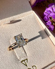 A radiant cut white diamond ring with a gold band displayed inside a pink jewelry box, with purple flowers in the background.