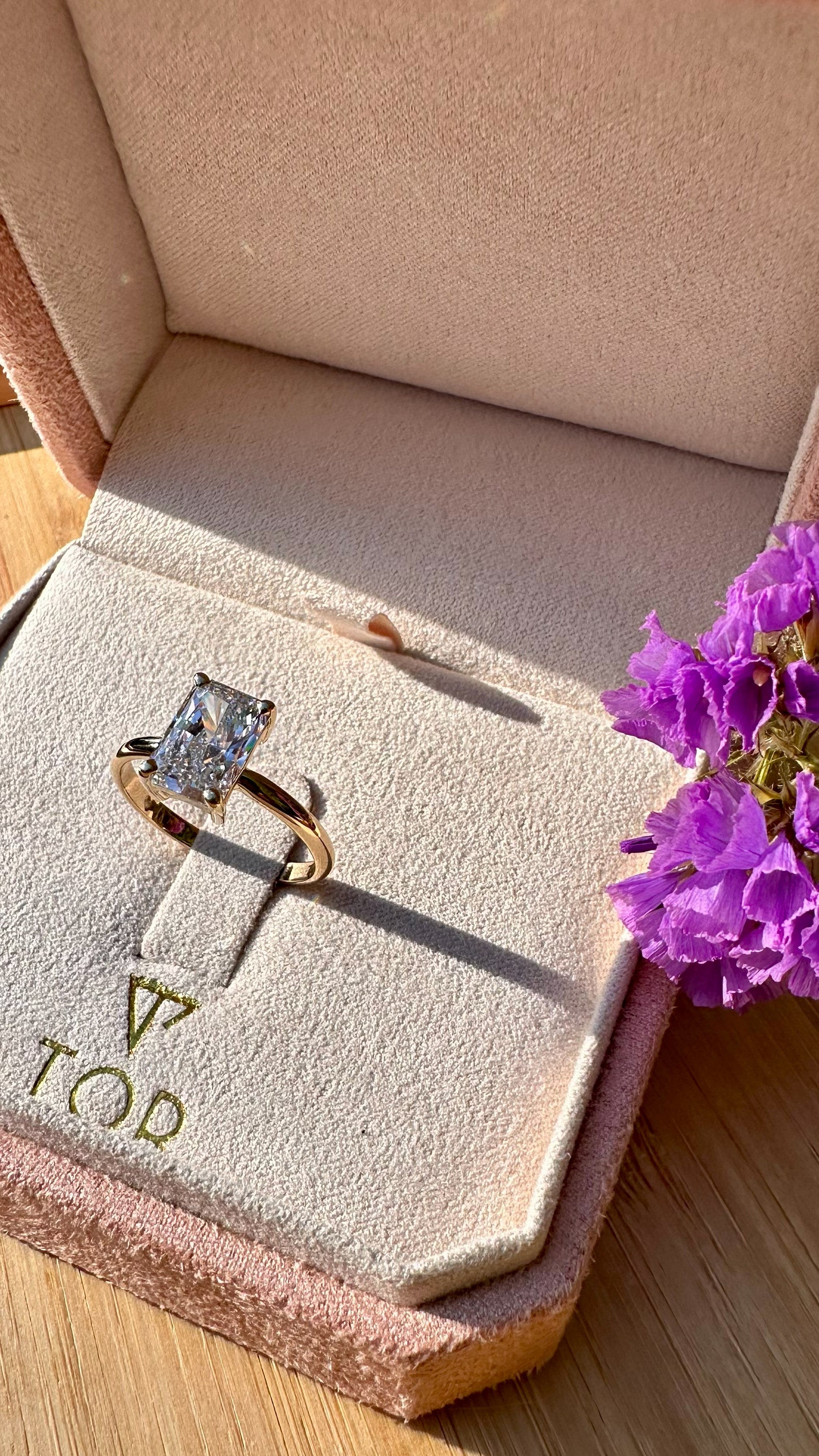A gold ring with a radiant cut white diamond displayed on a beige velvet ring box, set on a wooden surface with purple flowers nearby.