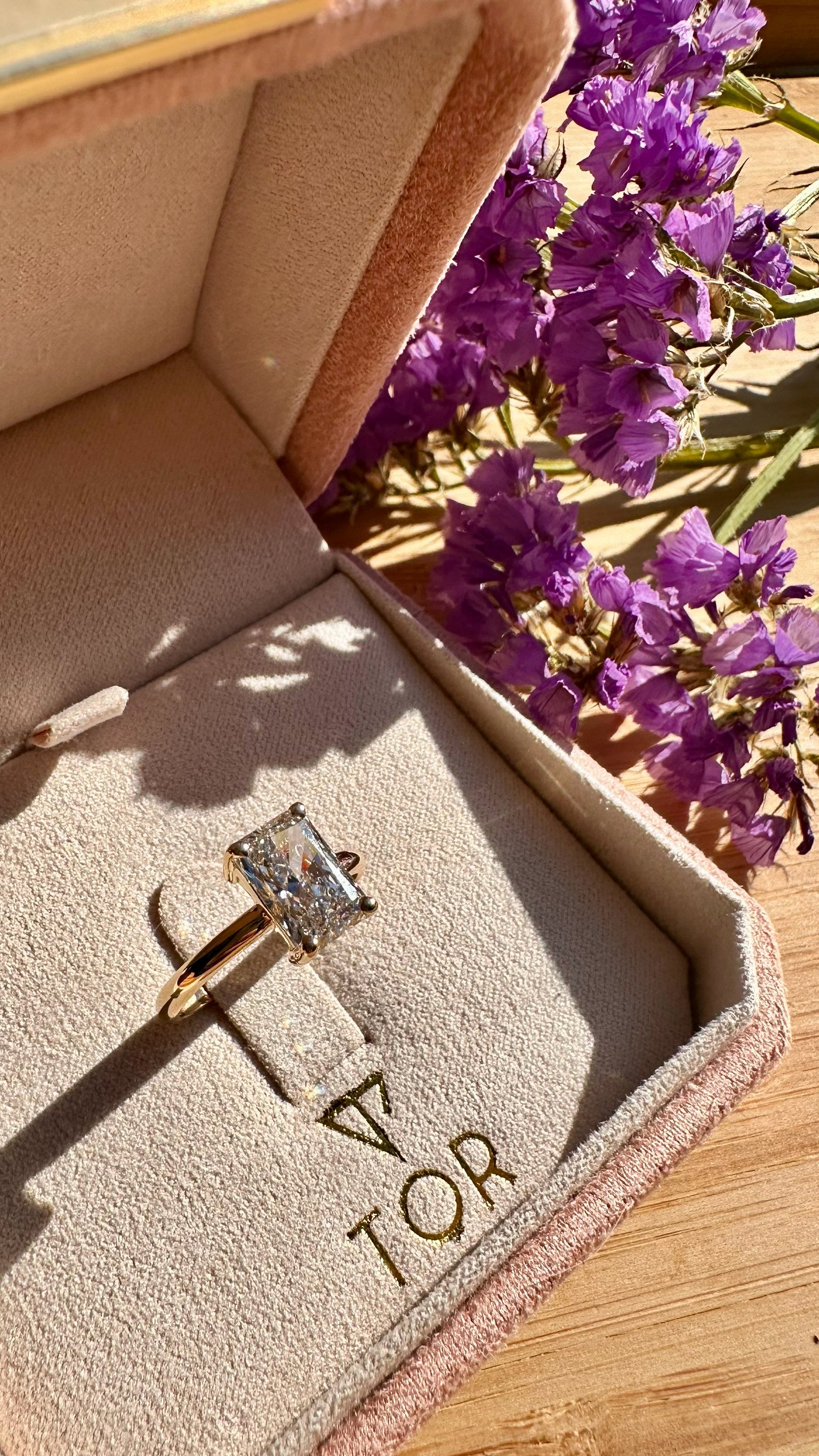 A gold ring with a radiant cut white diamond displayed inside a pink jewelry box, with purple flowers in the background on a wooden surface.