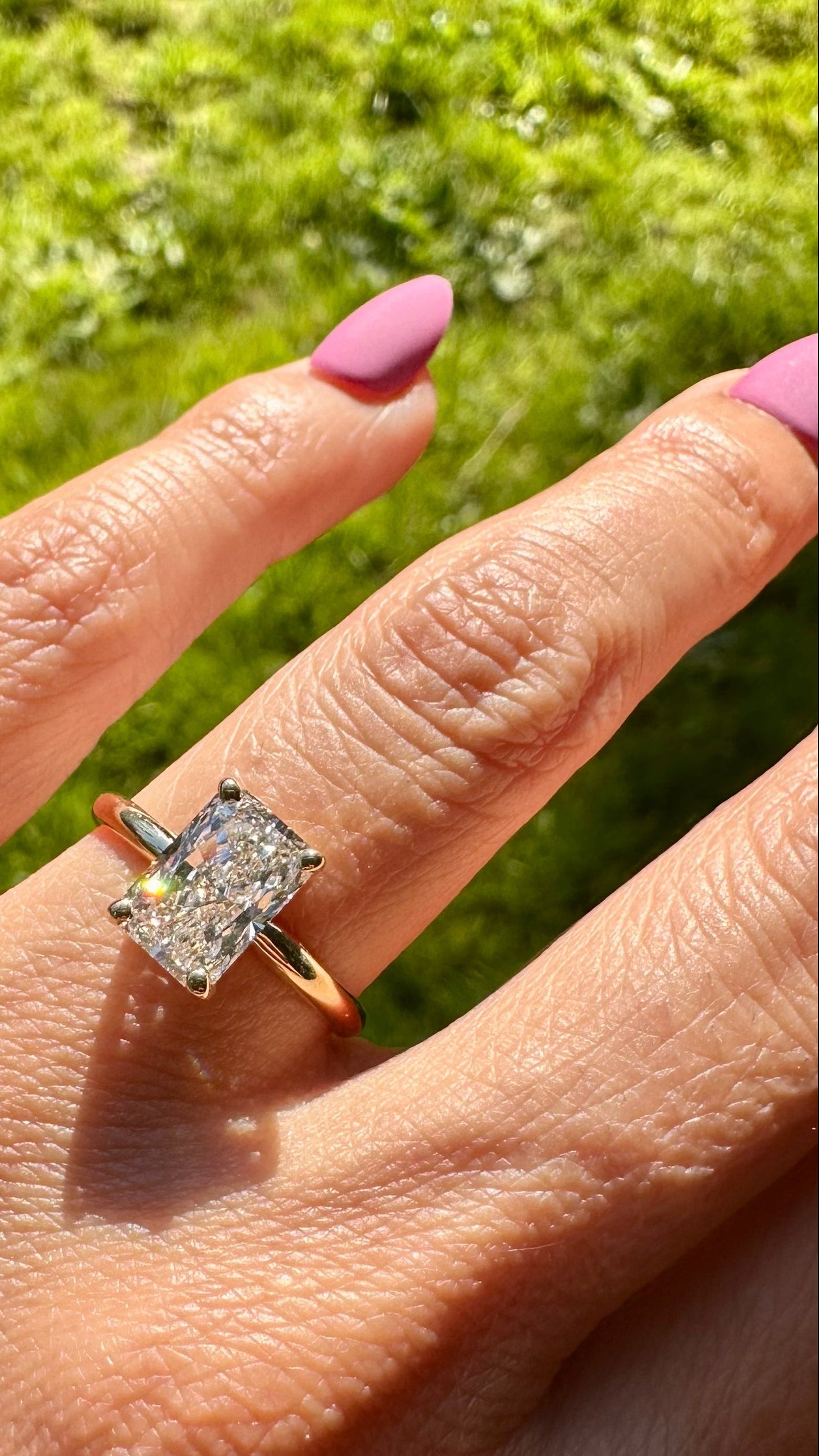 A gold ring with a radiant cut lab grown diamond worn on a person's ring finger, shown outdoors against a bright green grassy background.