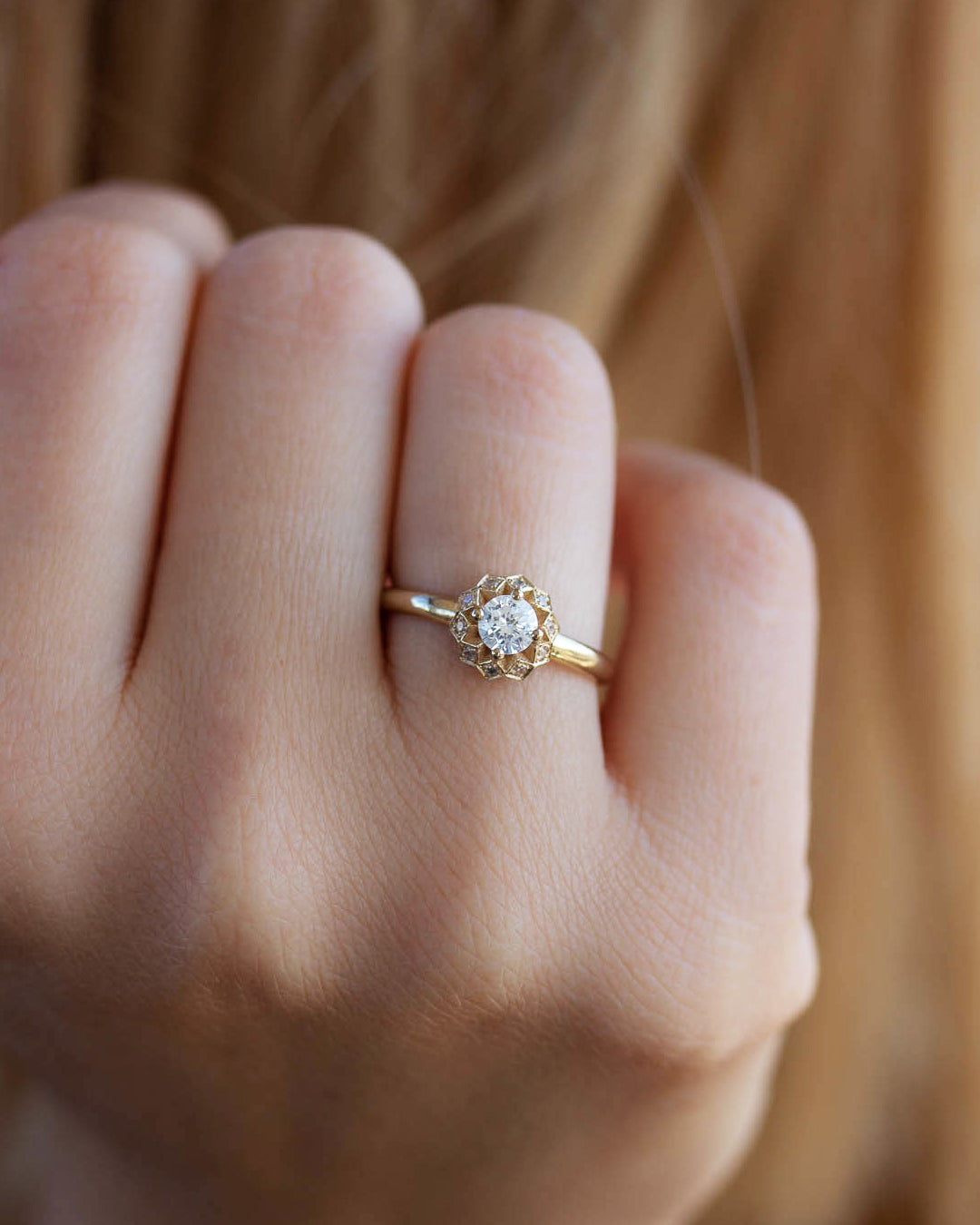 A delicate 14k yellow gold engagement ring, set with a center brilliant cut white diamond and smaller diamonds around it, in the shape of a geometric flower. 